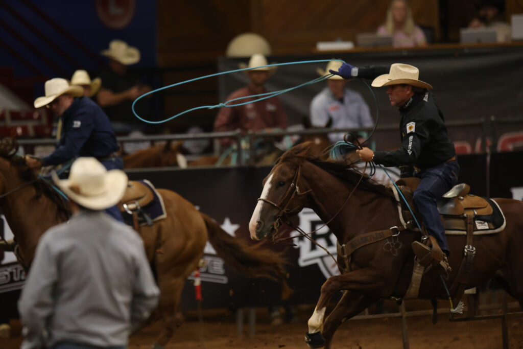 Cowboy on brown horse swings blue rope during indoor rodeo roping event, with other competitors and spectators visible in arena