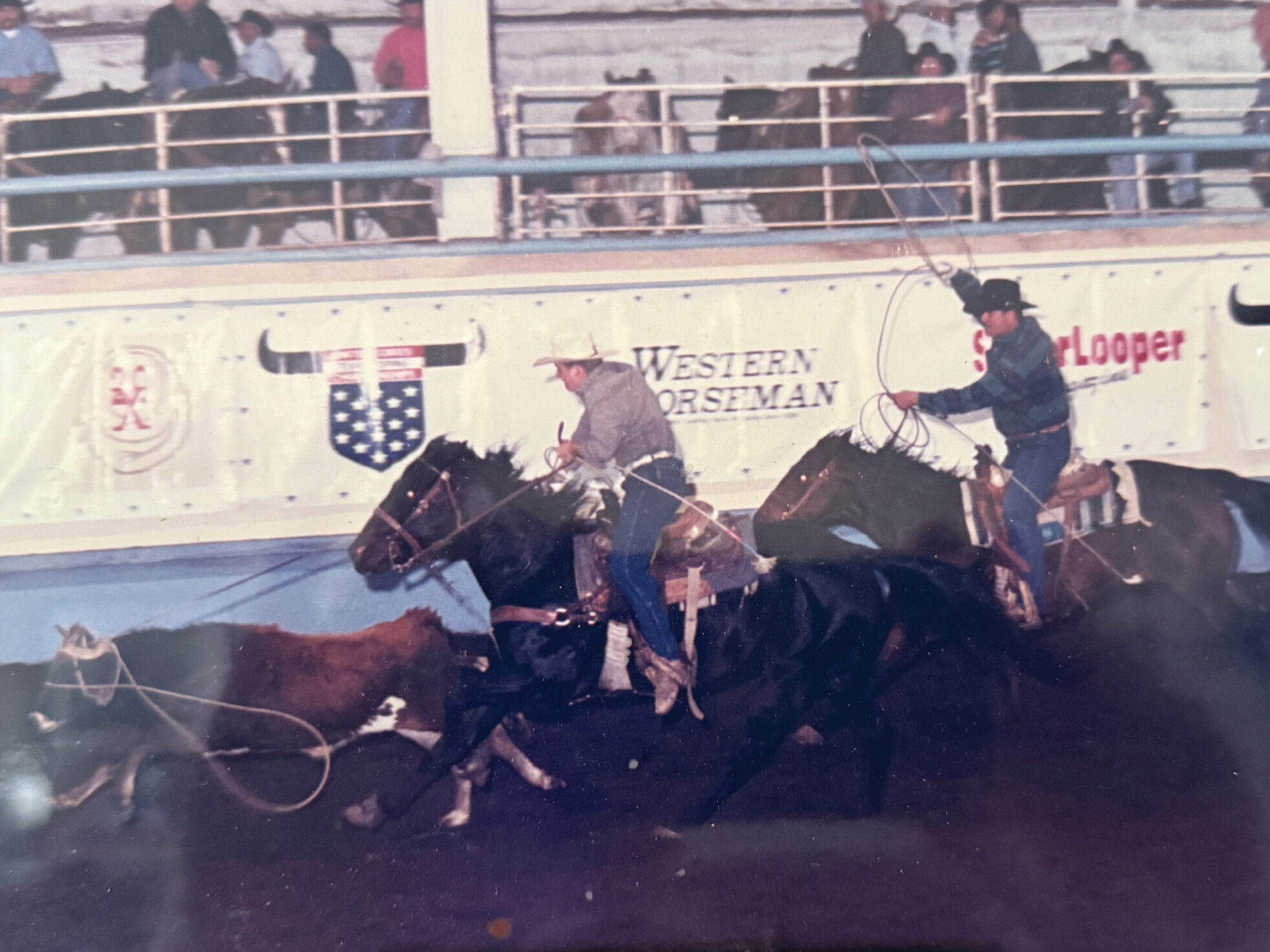 Cowboy on horseback swinging a lasso during a rodeo event at an arena with "Western Horseman" signage visible