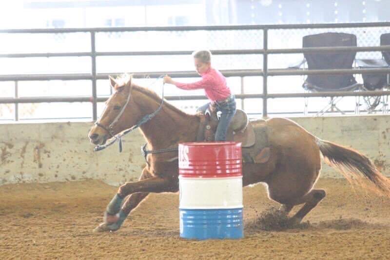 Young rider in pink shirt barrels around a red, white and blue drum on horseback during a rodeo event