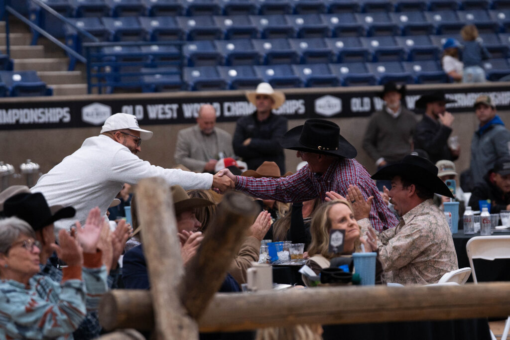 Man in white cap shakes hands with spectator in black cowboy hat at rodeo event in arena grandstand