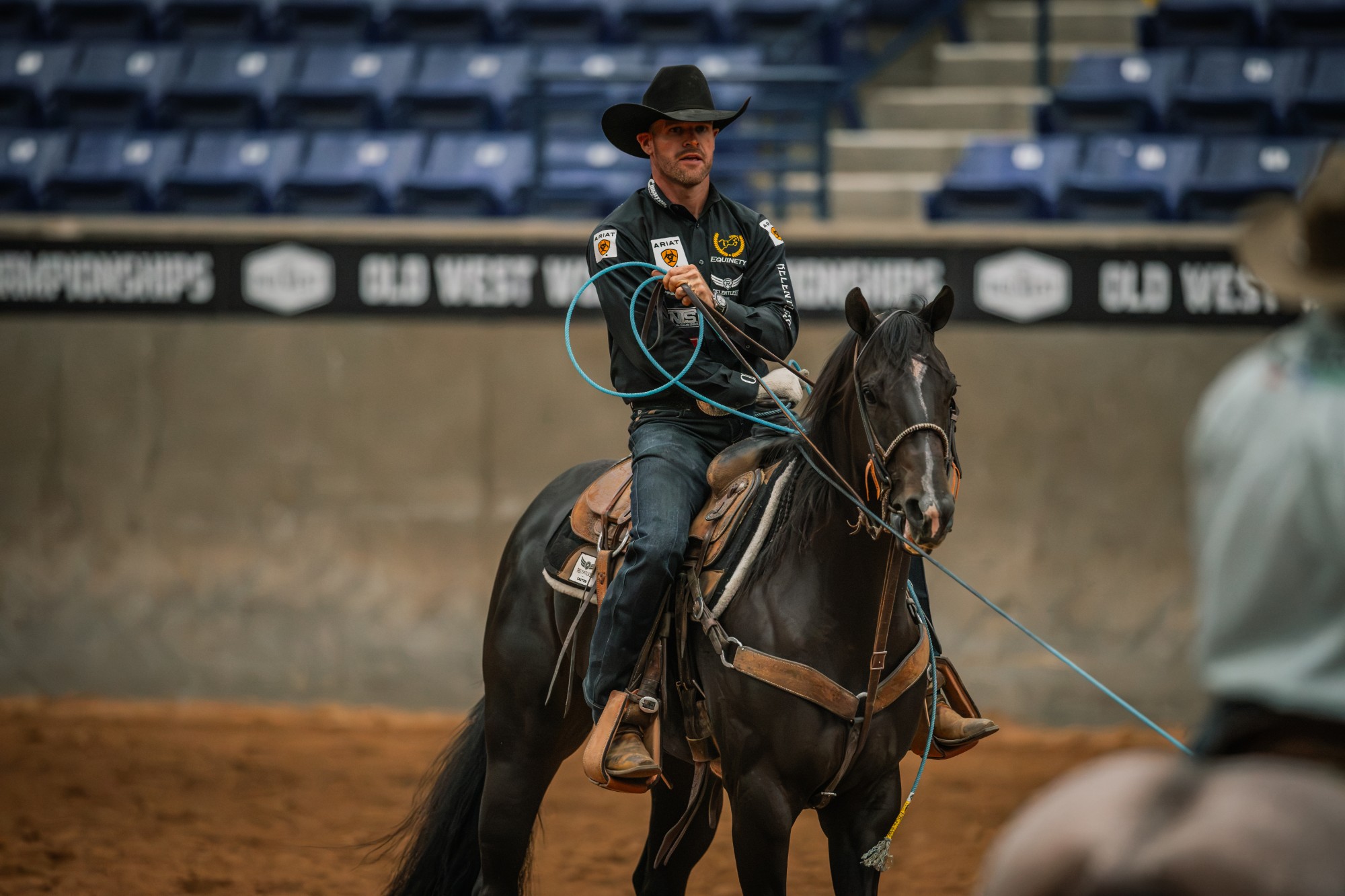 Miles Baker rides Fendiii toward the camera in an empty arena with Old West World Championships banners behind him.