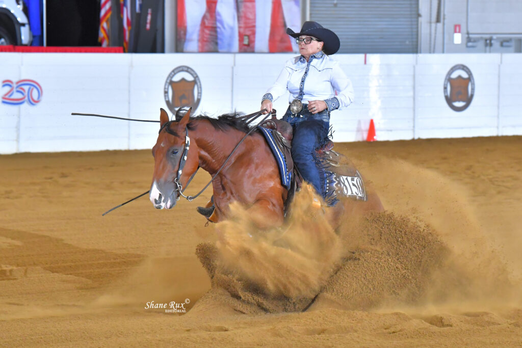Barrel racer on a brown horse kicks up dust while turning a barrel at an indoor rodeo arena
