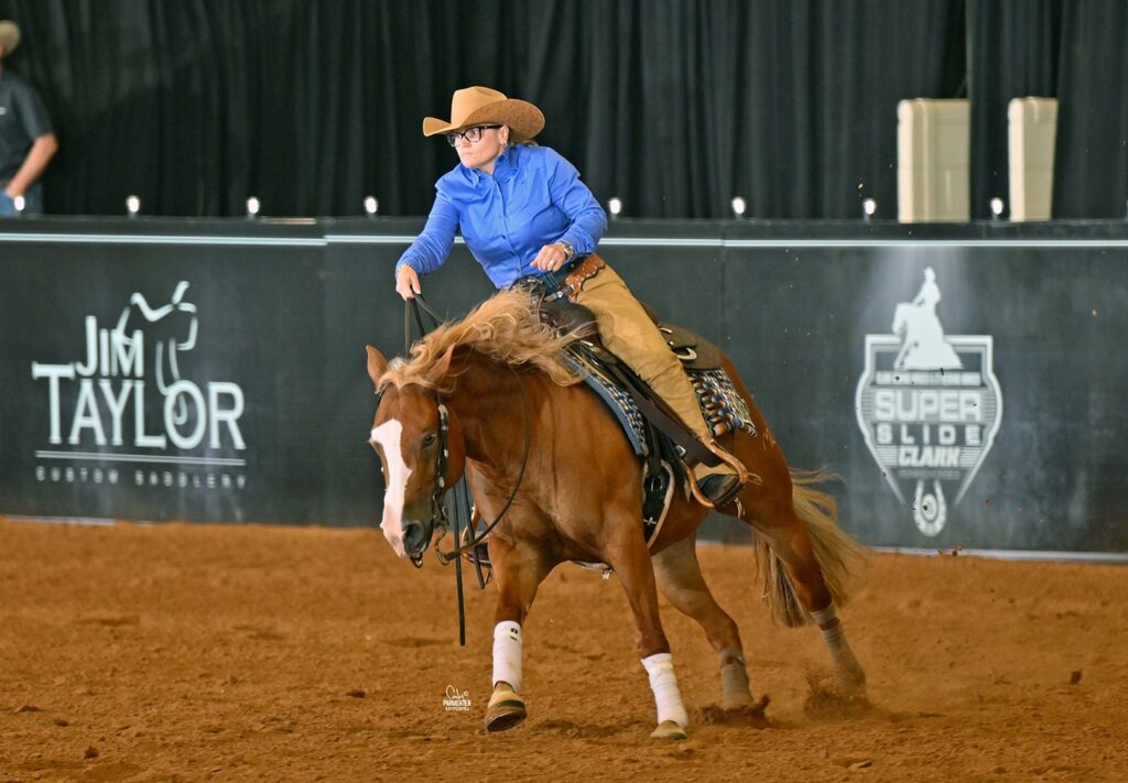Cowboy on brown horse with white blaze competing in reining event at indoor arena with Jim Taylor Saddles signage