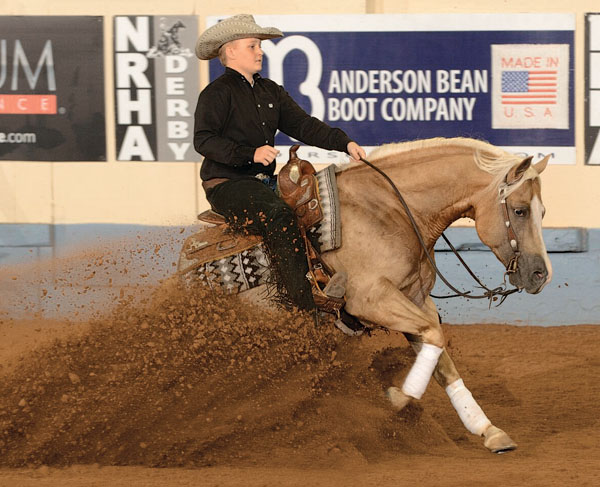 Cowboy in black shirt and tan hat performs sliding stop on palomino horse during reining event at indoor arena with Anderson Bean Boot Company signage