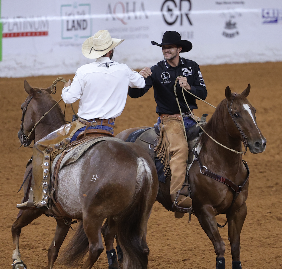 Two cowboys on horseback interact in a rodeo arena, one wearing a white shirt and cream cowboy hat, the other in black shirt and black cowboy hat