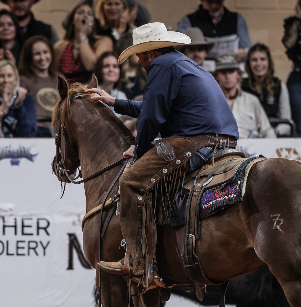 Cowboy in white hat and blue shirt riding brown horse adjusts horse's bridle before spectators at rodeo event