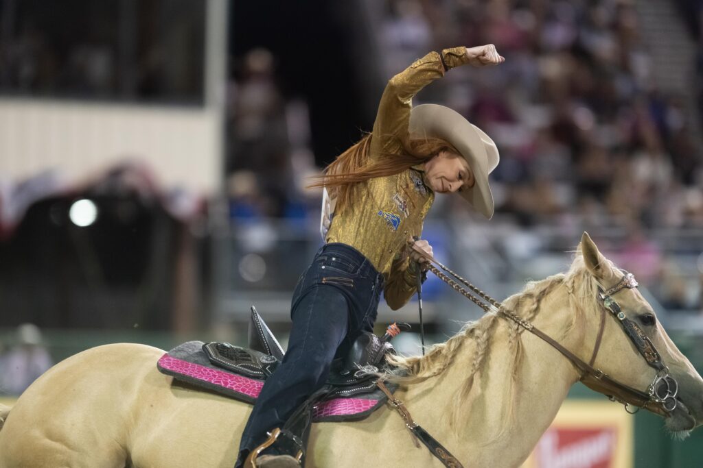 Cowgirl in a gold shirt and tan hat leans forward with arm raised while riding a palomino in a pink barrel racing saddle
