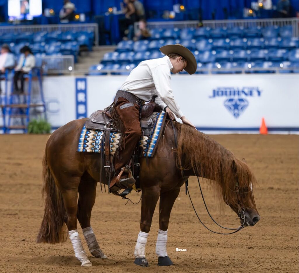 Cowboy on brown horse wearing white shirt and tan hat performs in indoor arena with blue seating and John Deere signage visible