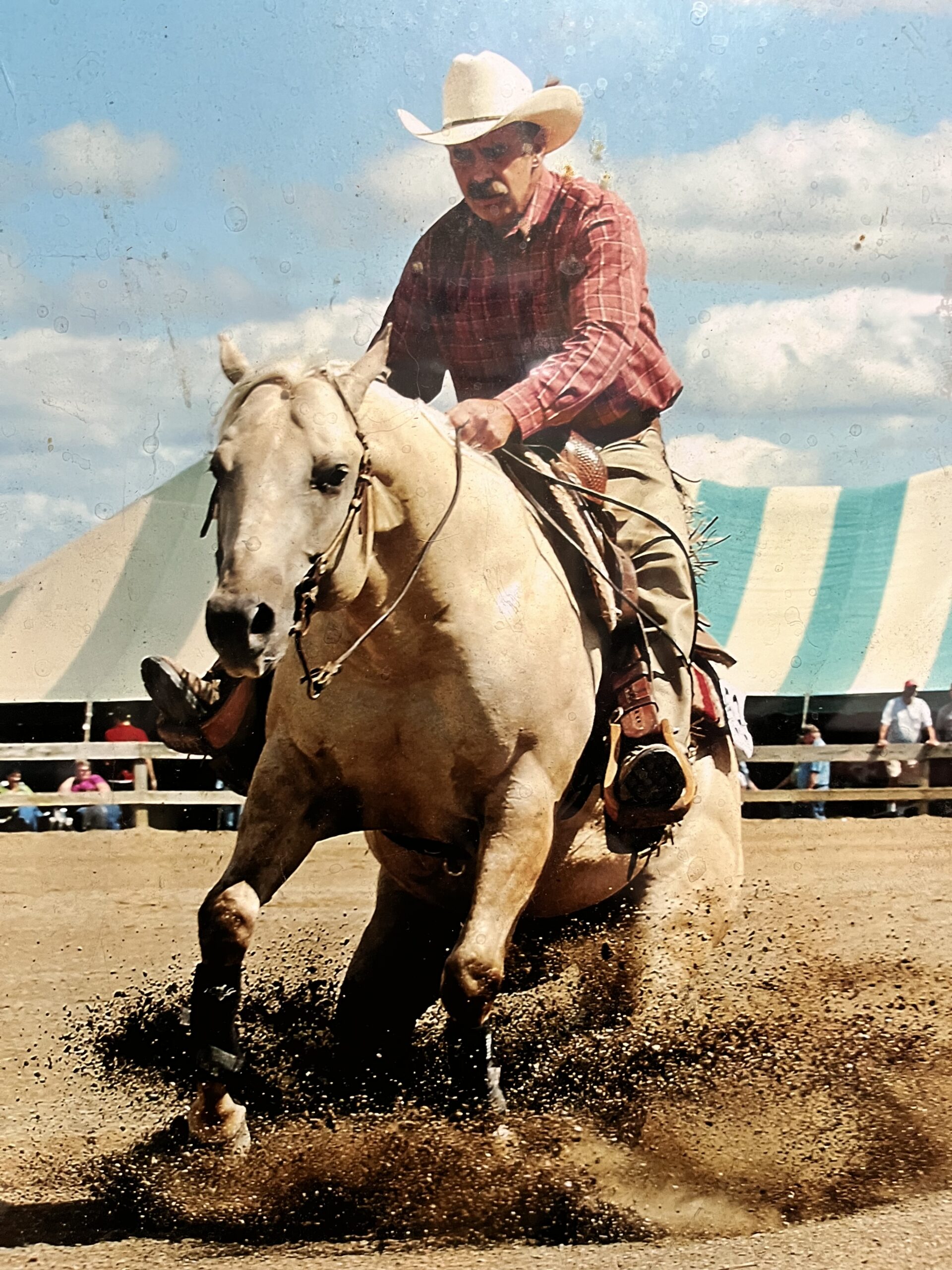 Cowboy in plaid shirt and tan hat rides white horse through arena dirt at rodeo with spectators in stands behind striped awning