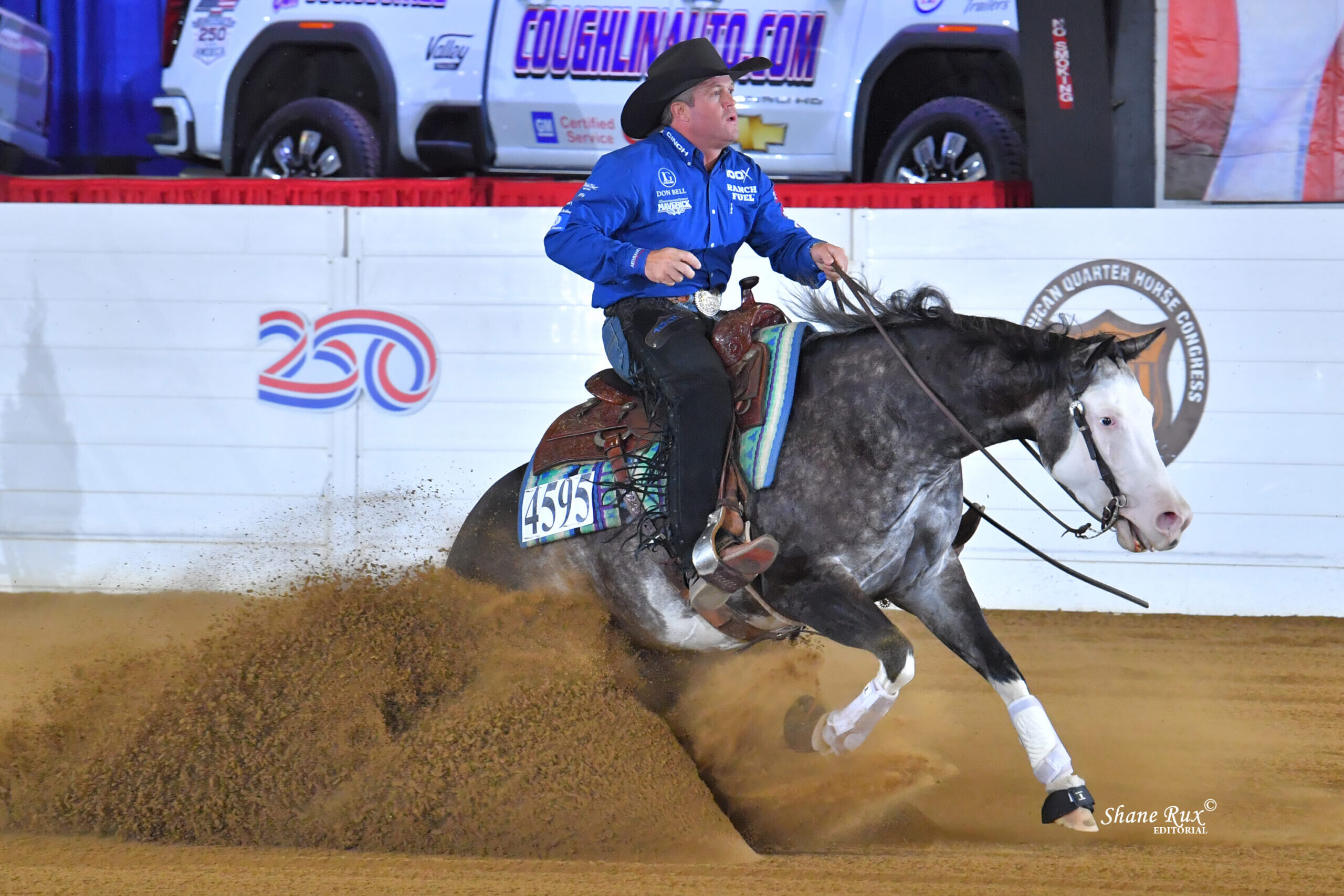 Cowboy in blue shirt and black hat rides gray and white horse sliding through dirt arena at indoor rodeo event
