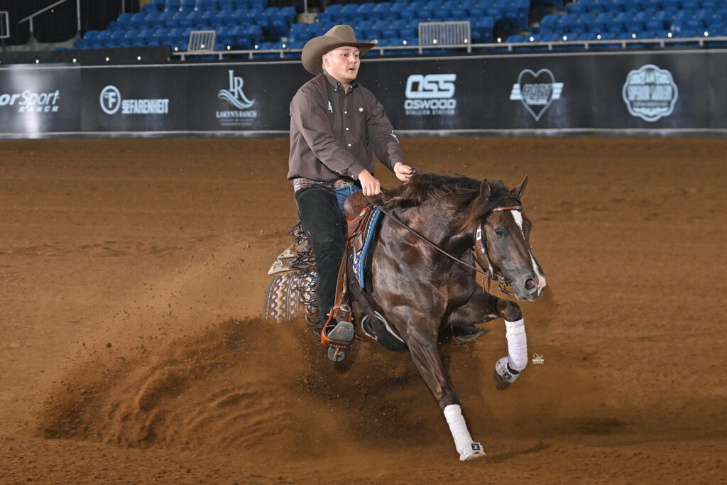Cowboy in brown shirt and tan hat rides a dark brown horse across a dirt arena at a professional rodeo event, kicking up dust
